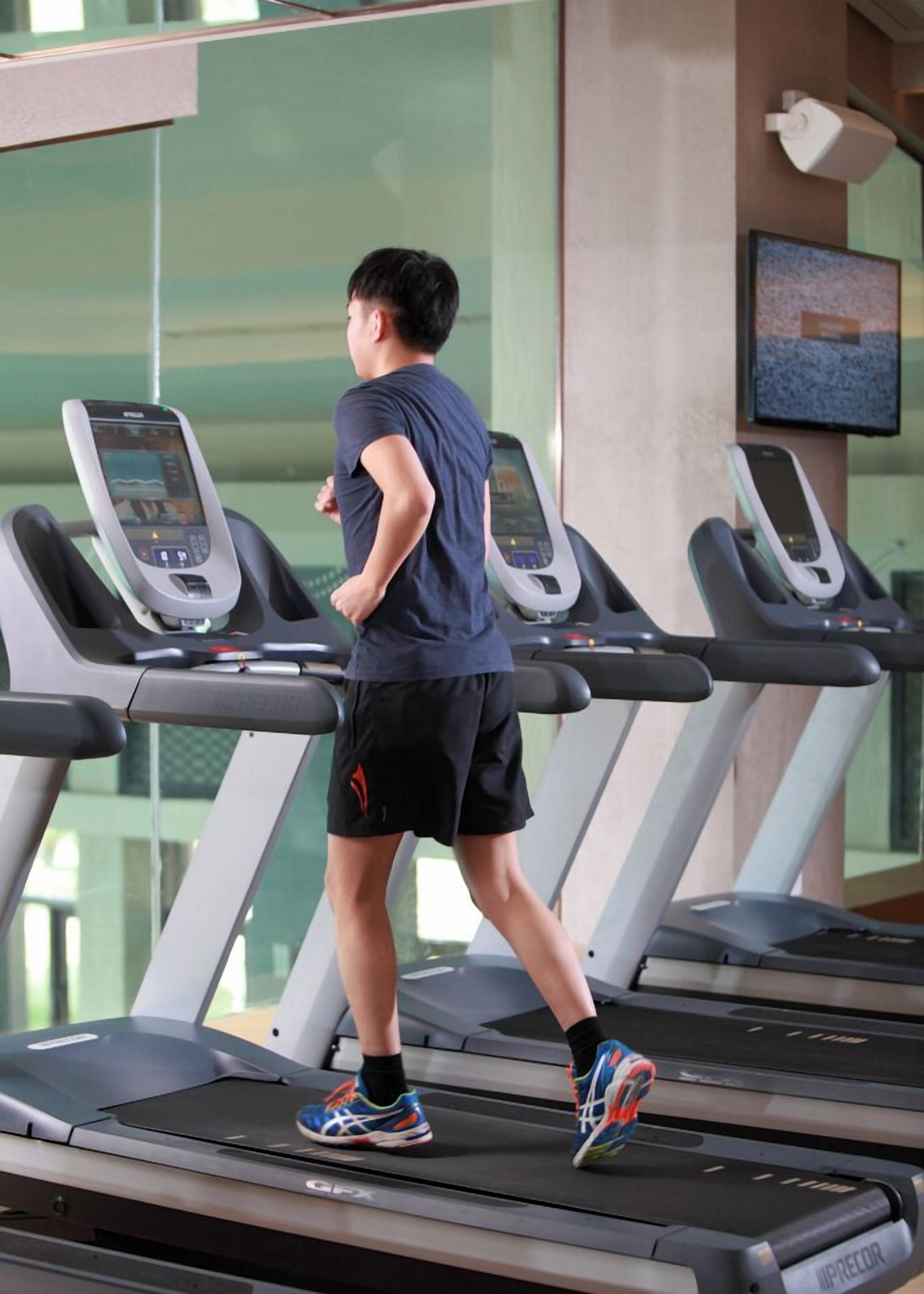 A person running on a treadmill in a gym with multiple treadmills, a large window, and a wall-mounted screen.