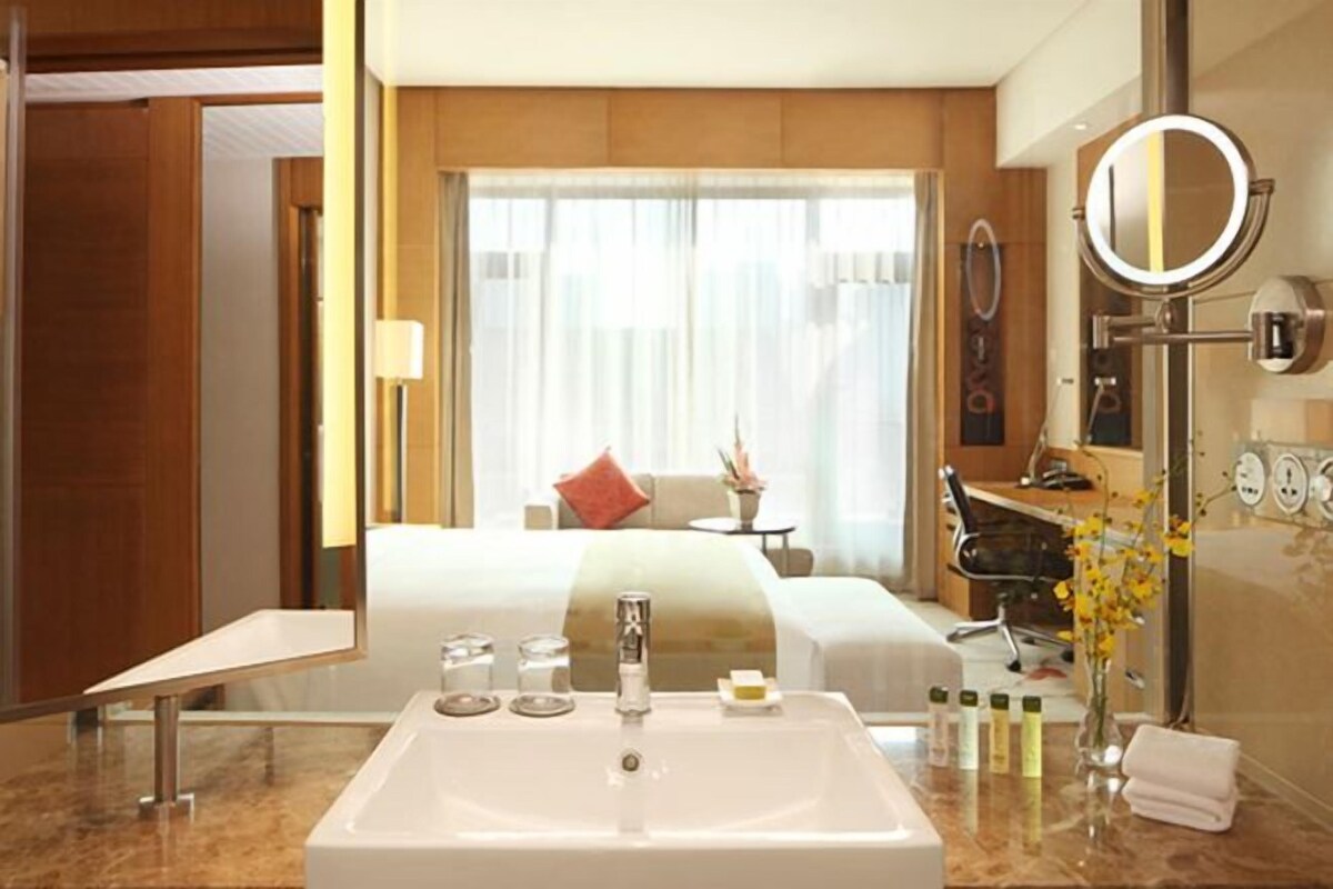 Un baño de hotel con un lavamanos grande, un espejo, artículos de aseo y vistas al dormitorio.