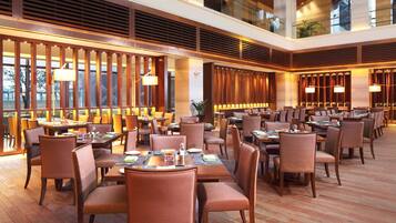 A spacious hotel lobby with multiple dining tables and chairs, large windows, and modern lighting fixtures.
