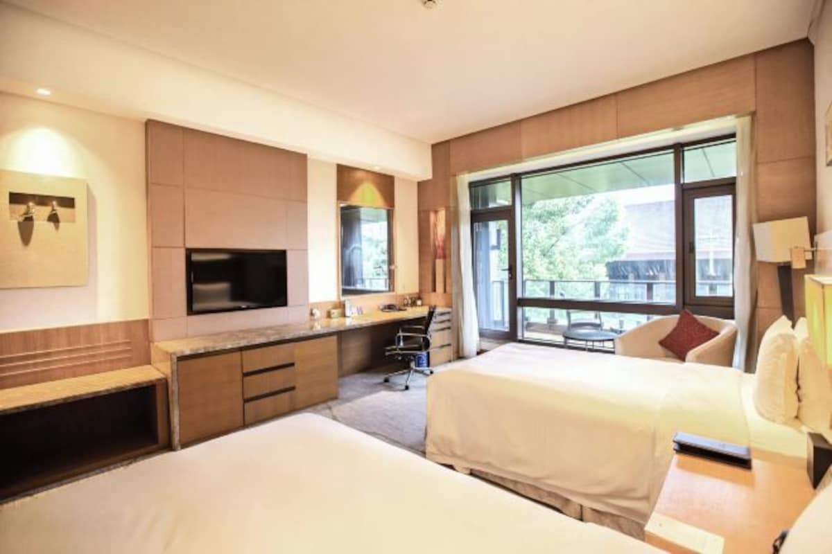 A hotel room with two beds, a desk, a chair, a TV, and a large window offering a view of the outdoors.