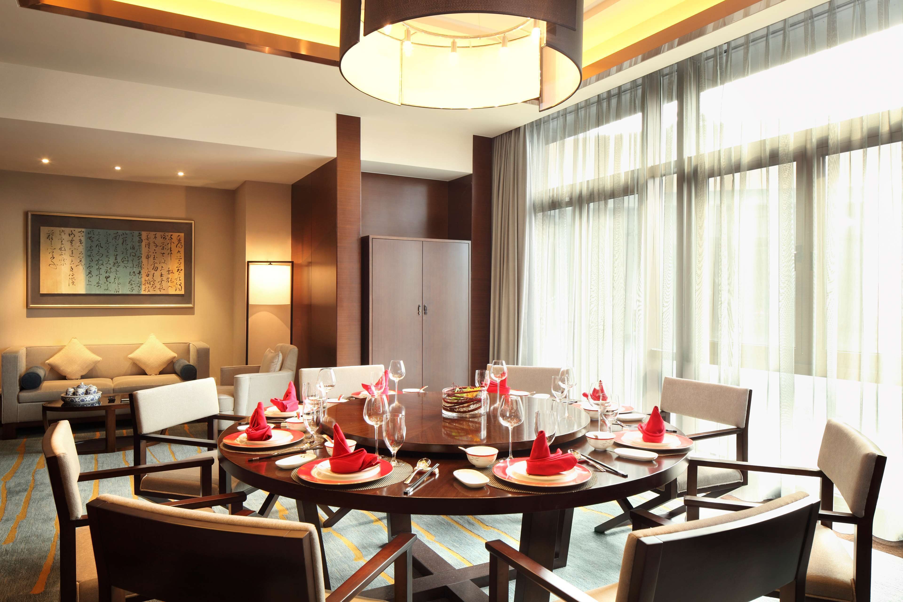 Une salle à manger d’hôtel avec une table ronde dressée pour un repas, entourée de chaises. On aperçoit un canapé et une lampe au fond, ainsi qu’une grande fenêtre avec des rideaux.
