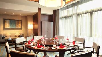 Une salle à manger d’hôtel avec une table ronde dressée pour un repas, entourée de chaises. On aperçoit un canapé et une lampe au fond, ainsi qu’une grande fenêtre avec des rideaux.