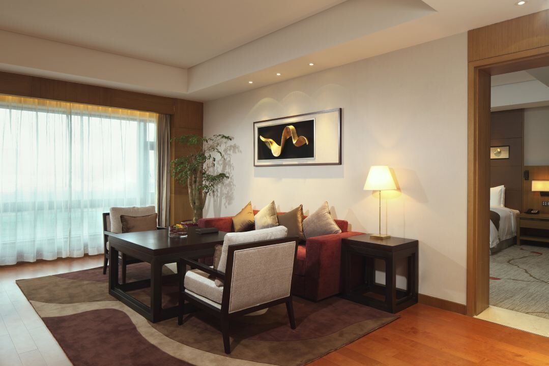 A modern living room with a dark wooden coffee table, a red sofa with multiple cushions, a beige armchair, and a floor lamp. There is a framed artwork on the wall and a large window with sheer curtains.