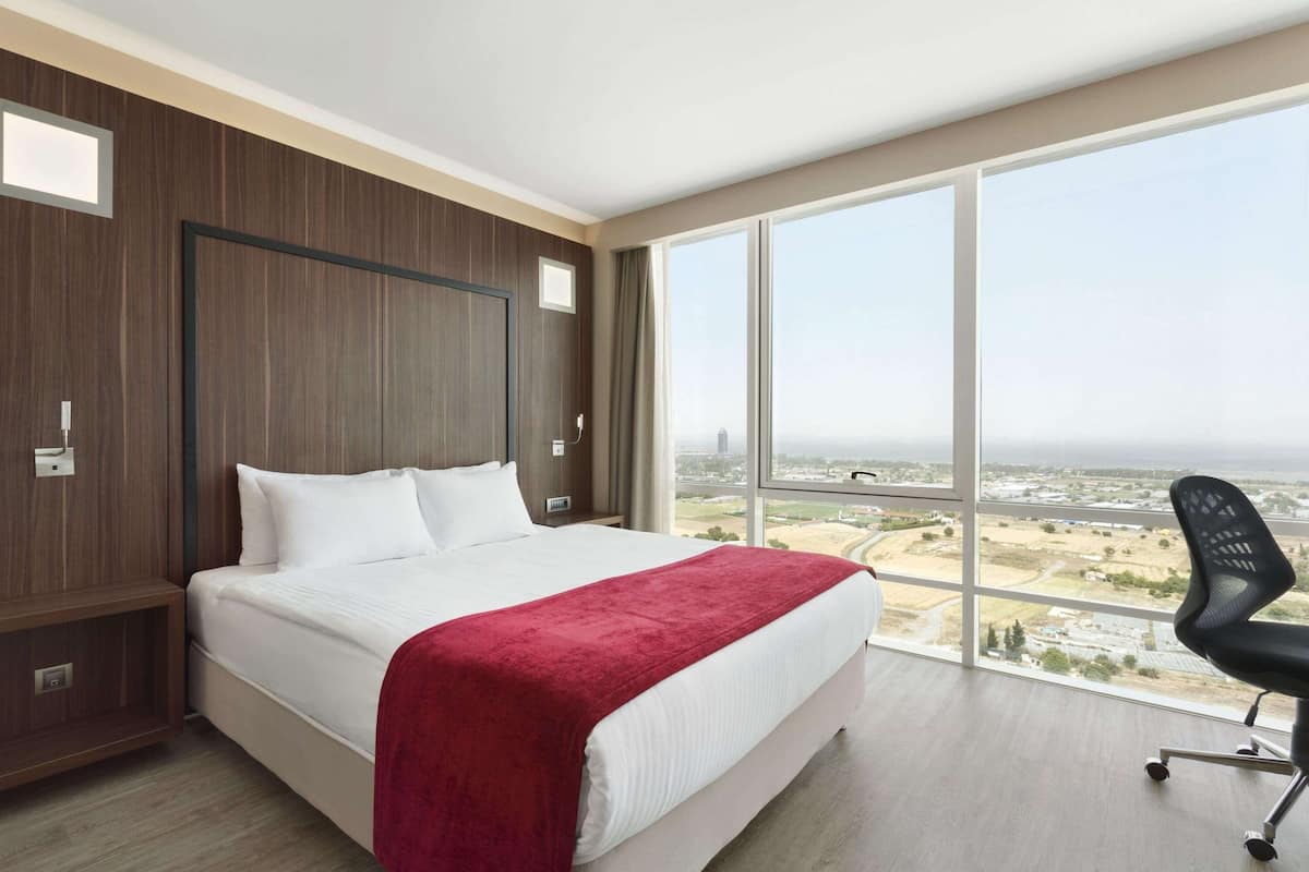 Quarto de hotel com uma cama grande, manta vermelha, cadeira de escritório preta e uma janela ampla com vista para a cidade.