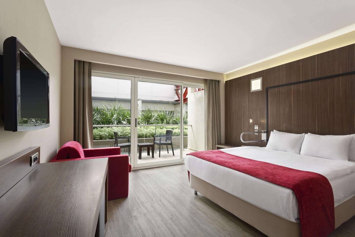 Quarto de hotel com uma cama grande, uma escrivaninha, televisão e varanda com mesa e cadeiras.