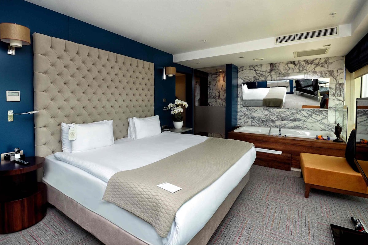 Quarto de hotel com uma cama grande, mesa de cabeceira, espelho, banheiro com banheira e parede de mármore.