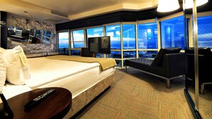 A hotel room with a large bed, a TV, a sofa, and a view of the cityscape through large windows.