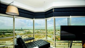 A modern room with a large window offering a city view, a black leather sofa, a white table, and a flat-screen TV.