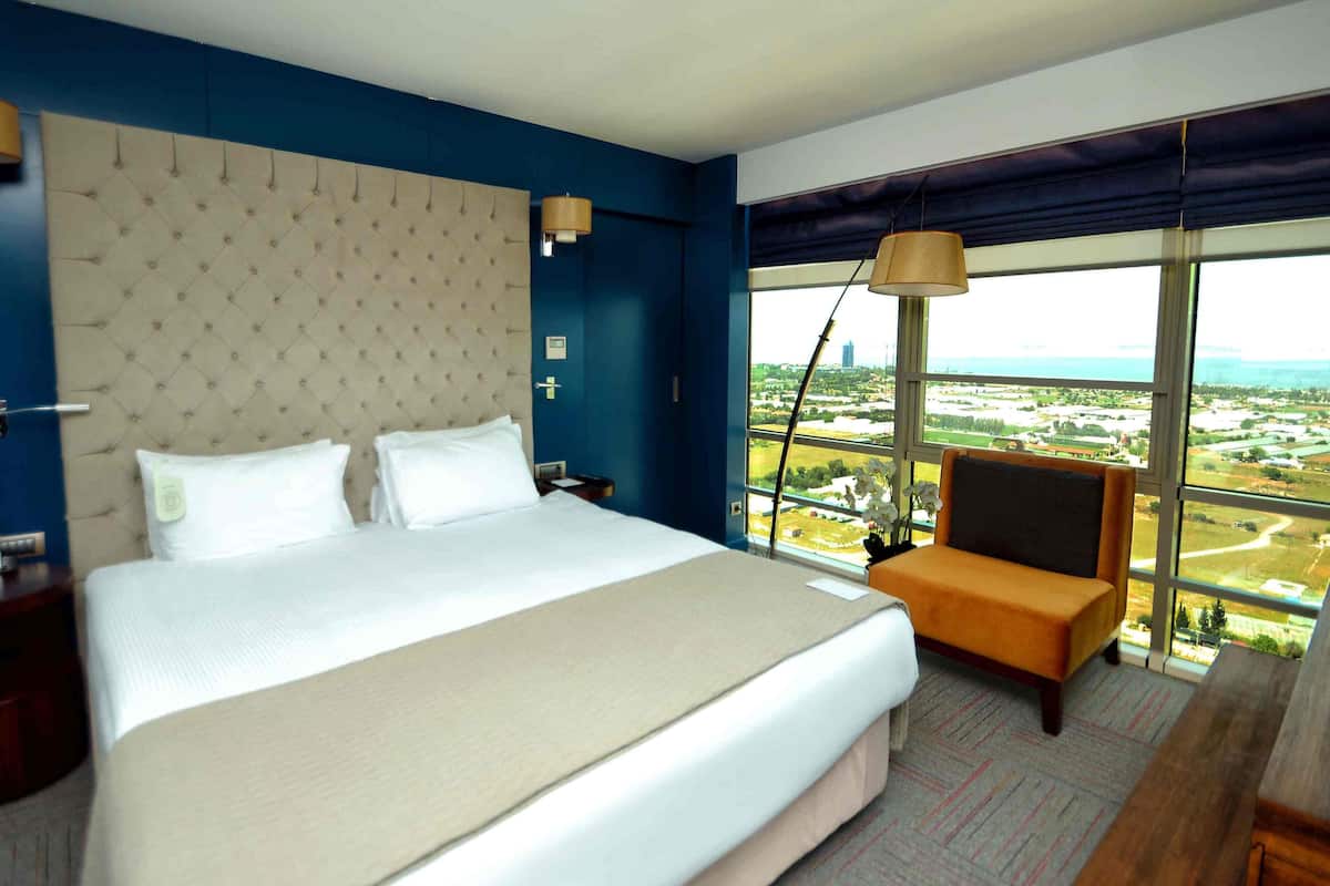 Quarto de hotel com uma cama grande, uma mesa de cabeceira, uma cadeira, uma janela ampla com vista e um abajur.