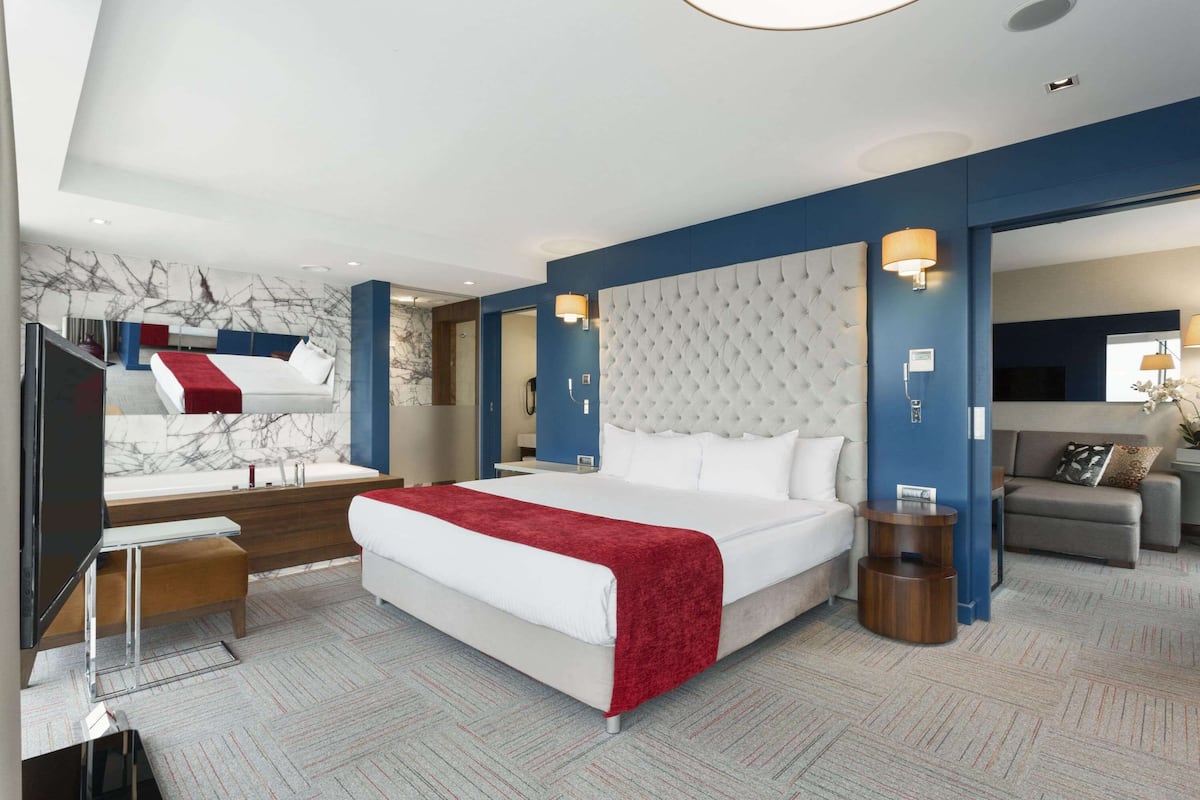 Quarto de hotel moderno com uma cama grande, televisão, escrivaninha e área de estar com sofá e poltrona.