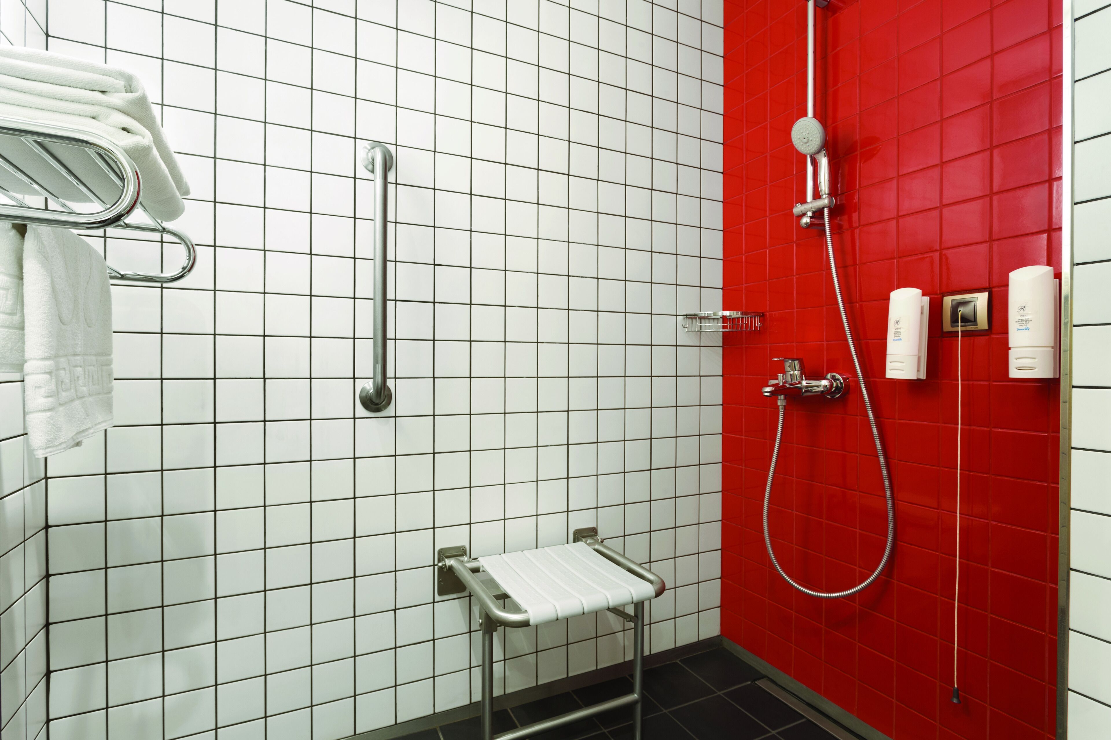 A bathroom with white tiled walls, a red accent wall, a handrail, a showerhead, and a towel rack.
