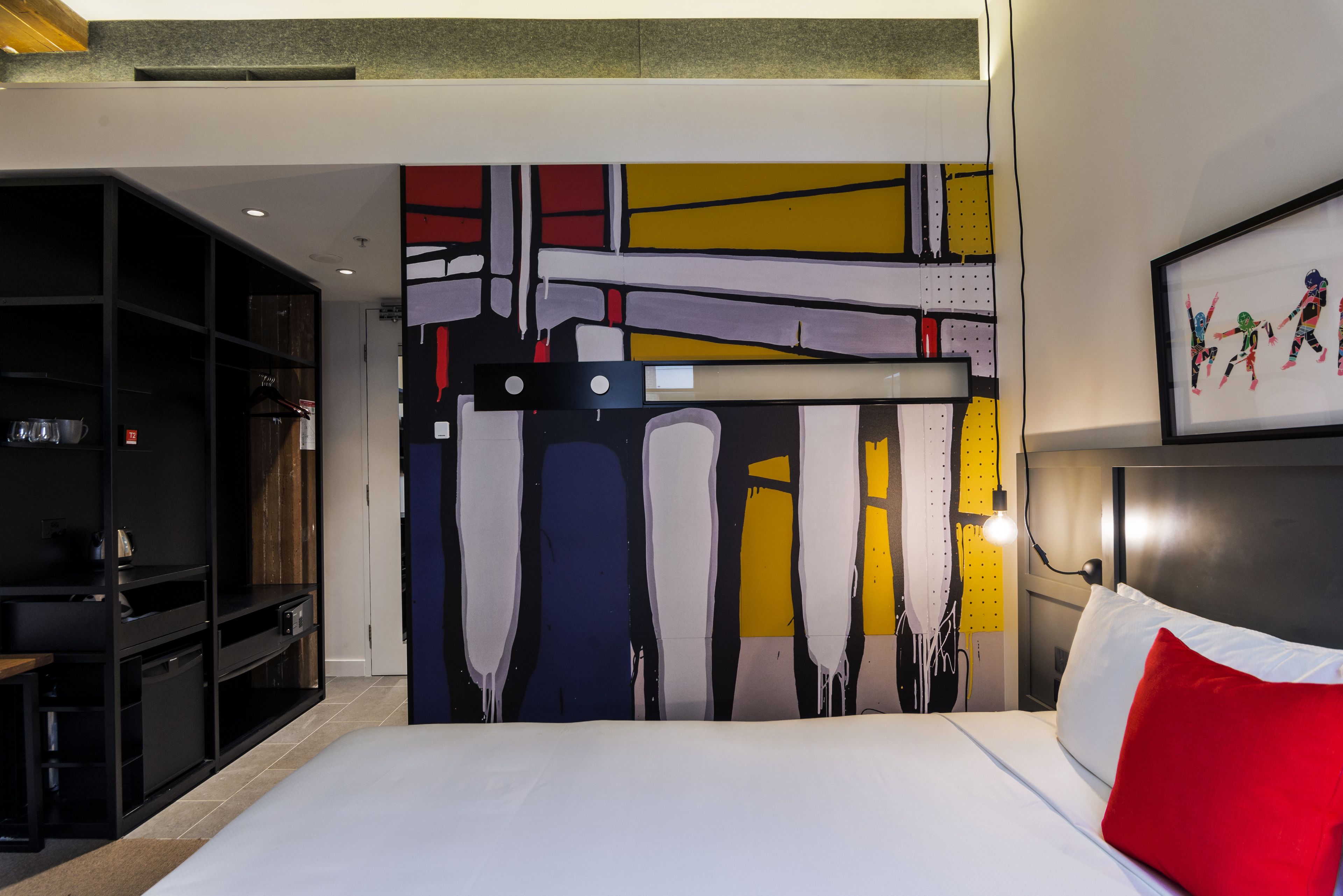 Une chambre d’hôtel avec un grand tableau abstrait au mur, un lit avec du linge de maison blanc et un oreiller rouge, et un bureau avec une chaise.