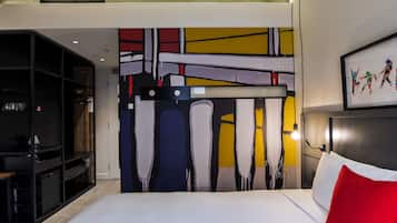 Une chambre d’hôtel avec un grand tableau abstrait au mur, un lit avec du linge de maison blanc et un oreiller rouge, et un bureau avec une chaise.