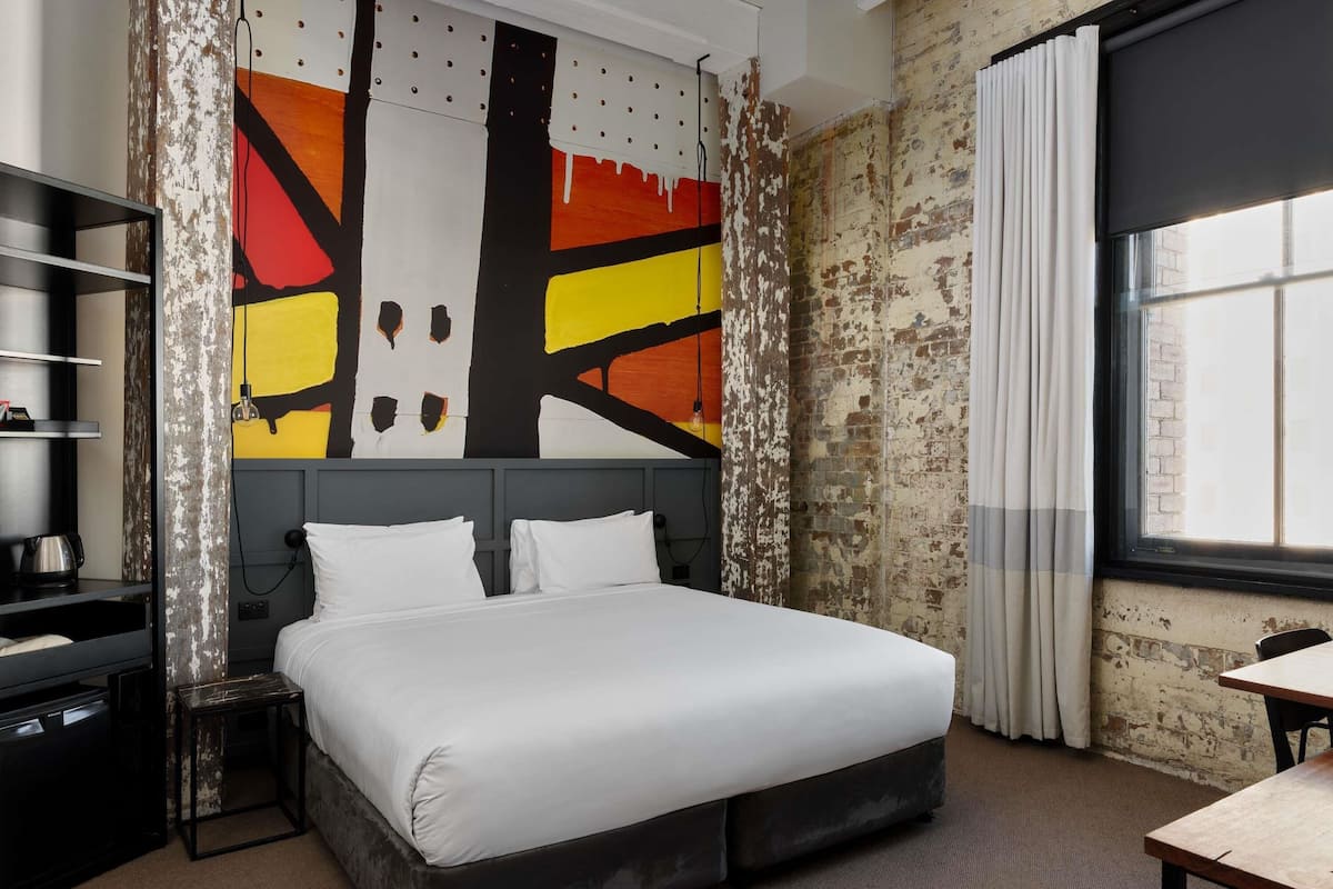 Habitación de hotel con cama, escritorio, silla, estantería y un cuadro abstracto grande en la pared.