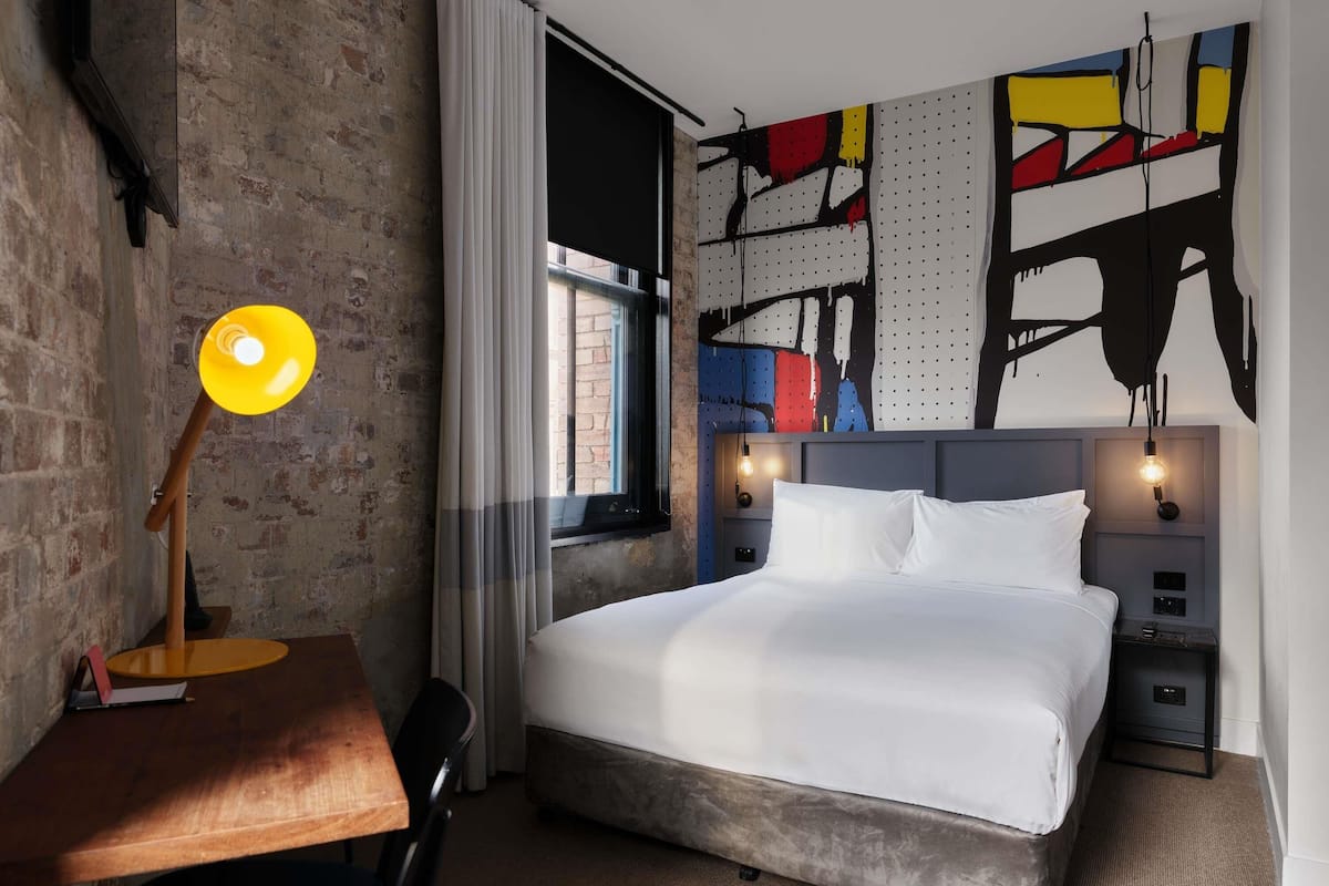 Una habitación de hotel moderna con una cama grande, un escritorio con lámpara y un cuadro abstracto y colorido en la pared.
