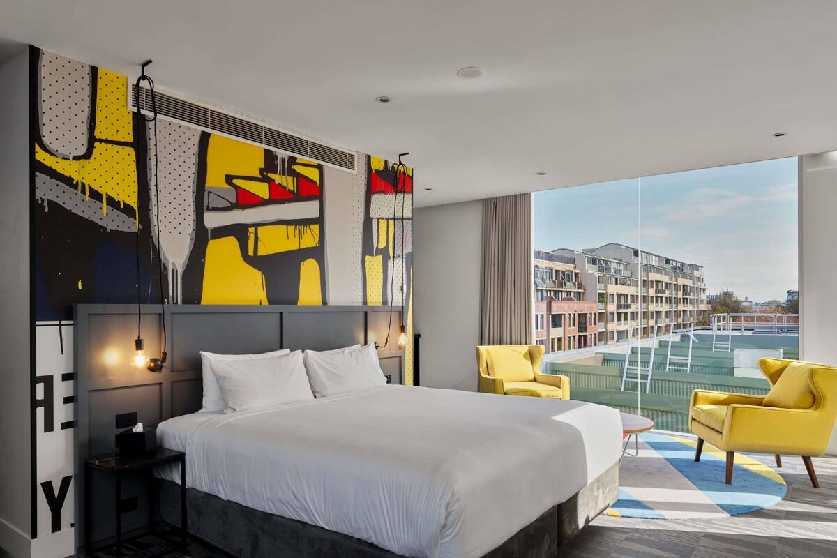A modern hotel room with a large bed, a view of the city, and a balcony with yellow chairs.