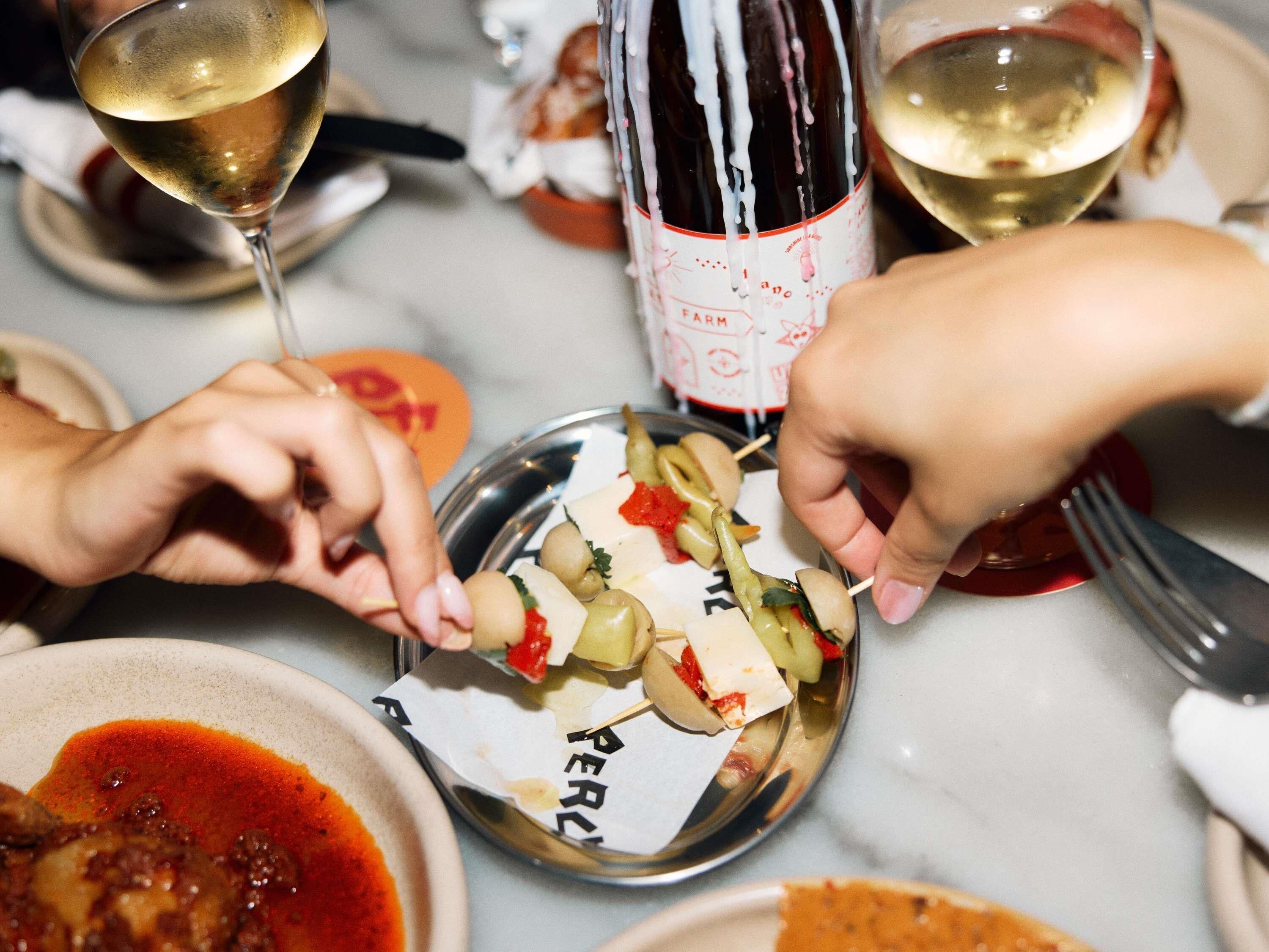 A dining table with a bottle of wine, glasses, and skewers of food.