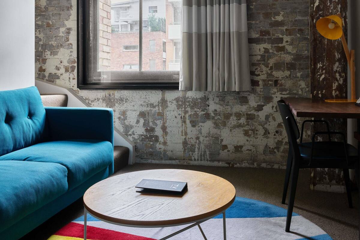 Una habitación con un sofá azul, una mesa de centro redonda de madera, una mesita con una silla y una ventana con cortinas.