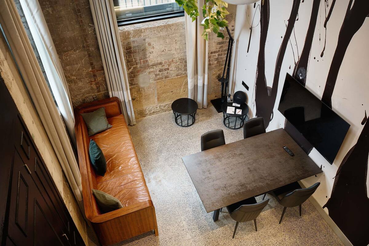 A cozy room with a brown leather sofa, a small round table, and chairs, a potted plant, and a TV mounted on the wall.