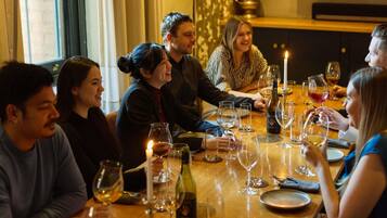 A group of people seated around a wooden dining table with wine glasses and a candle, in a room with a television and a brick wall.