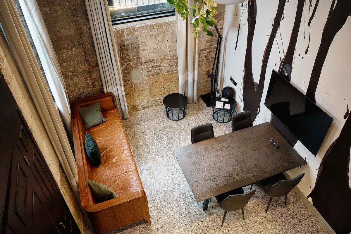 Una habitación acogedora con un sofá de cuero marrón, una pequeña mesa redonda y sillas, una planta en maceta y un televisor montado en la pared.