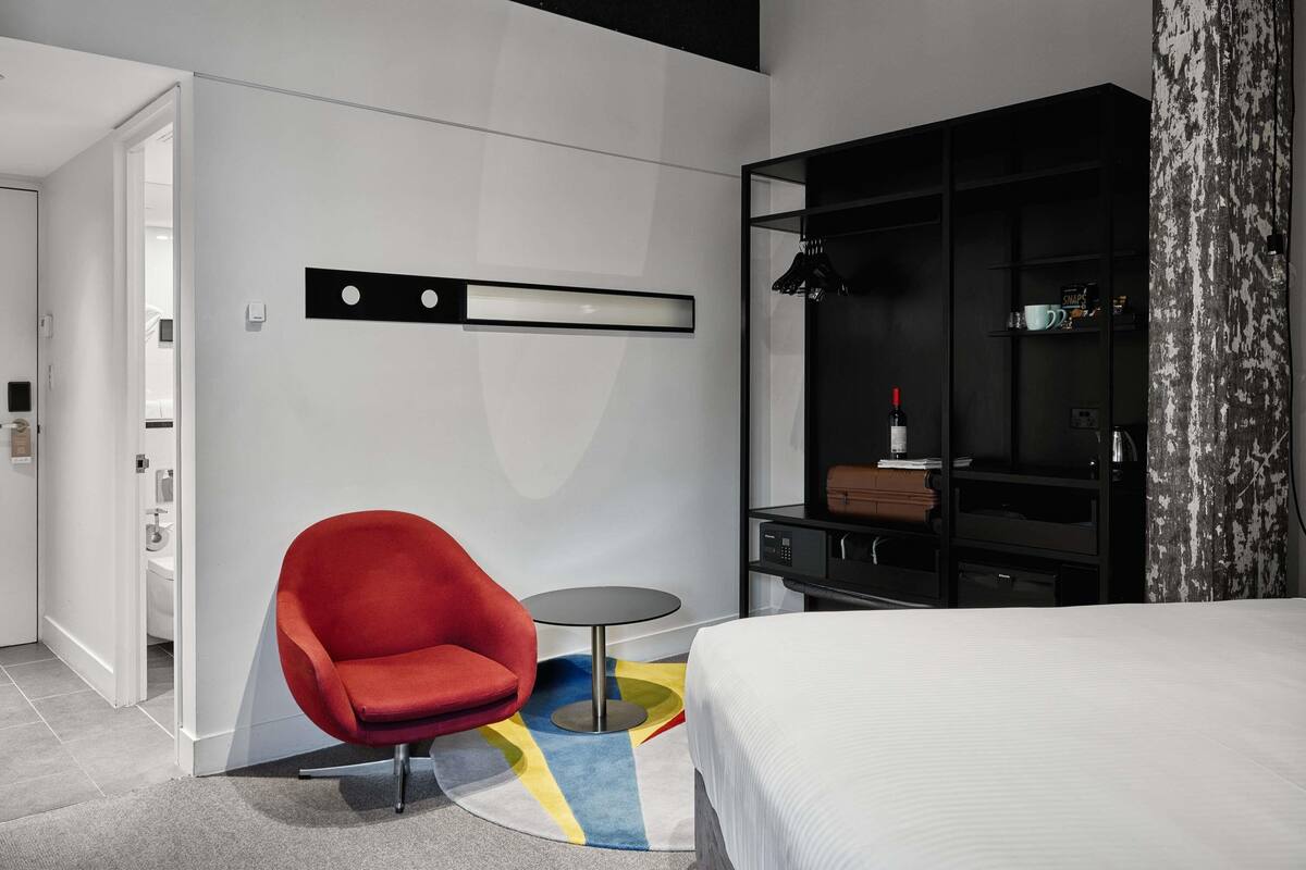 A modern hotel room with a bed, a red armchair, a small round table, a black wardrobe, and a white wall with a black shelf.