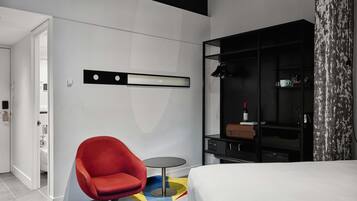A modern hotel room with a bed, a red armchair, a small round table, a black wardrobe, and a white wall with a black shelf.
