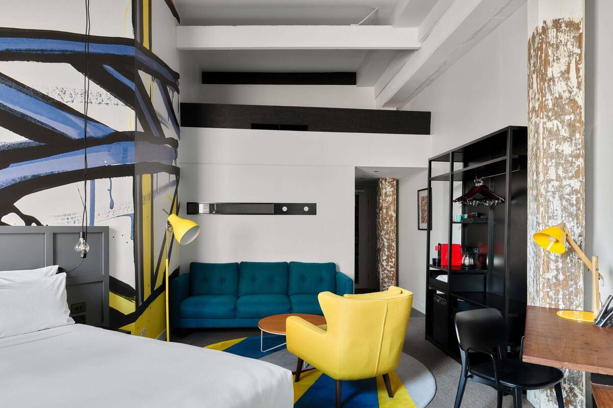 Una habitación de hotel moderna con una cama, un sofá, un sillón amarillo, un escritorio y una estantería.