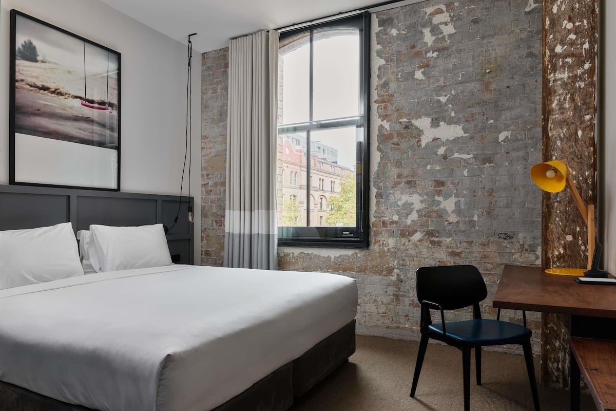 Ein Hotelzimmer mit einem Bett, einem Schreibtisch mit Stuhl, einer Lampe und Blick auf Gebäude durch ein Fenster.