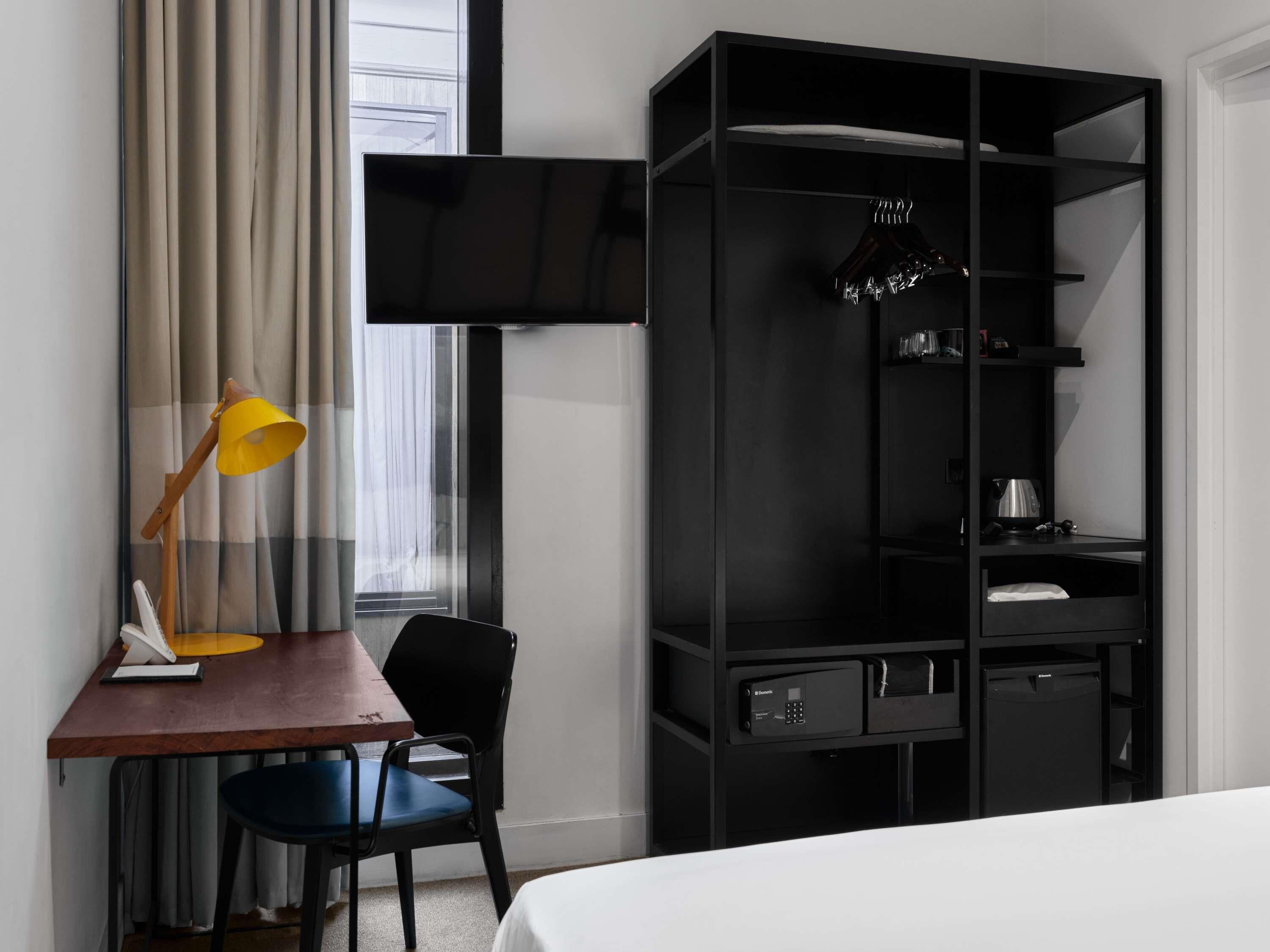 Habitación de hotel con cama, escritorio, silla, armario negro, televisor y ventana con cortinas.