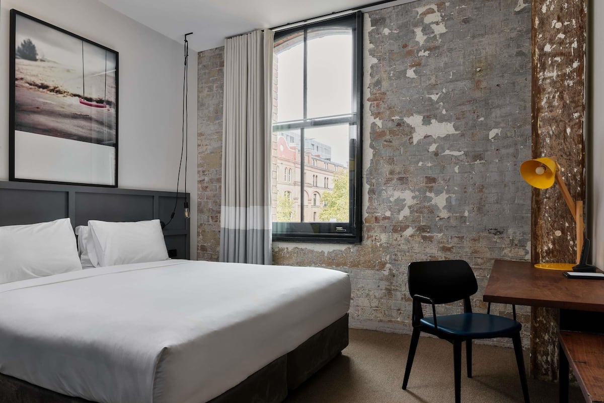 Habitación de hotel con cama, escritorio, silla, lámpara y vista a edificios a través de una ventana.