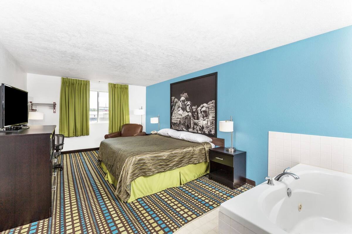 A hotel room with a bed, a desk, a TV, a bathroom with a bathtub, and a large window with curtains.