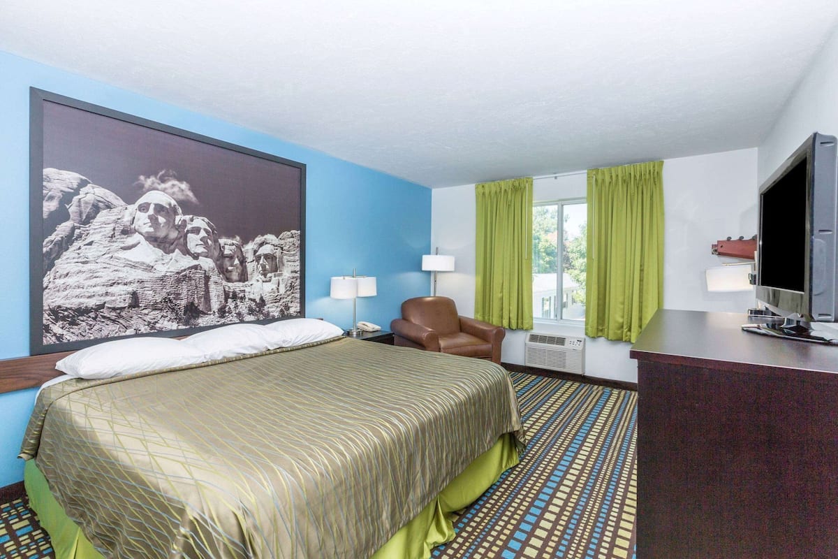 A hotel room with a bed, a chair, a desk with a TV, a large wall art, and a window with curtains.