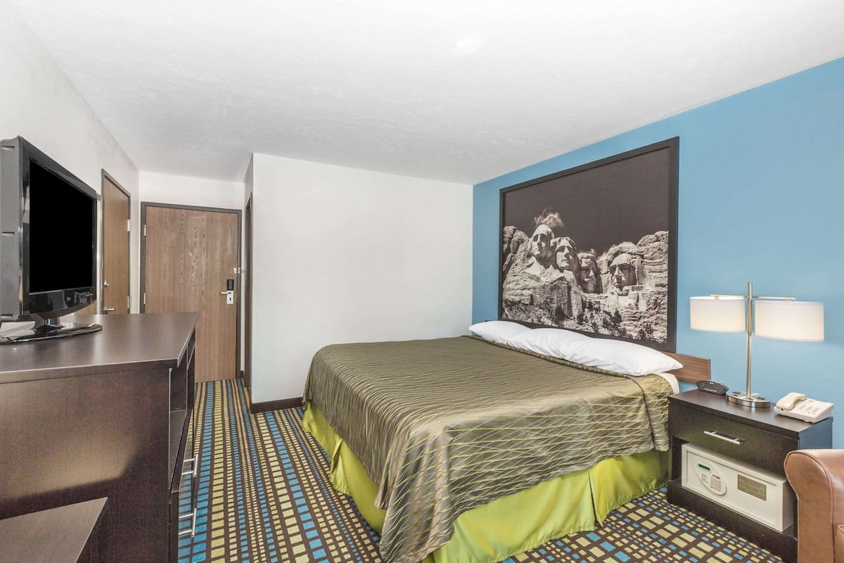A hotel room with a bed, a desk with a television, a nightstand with a lamp, and a patterned floor.