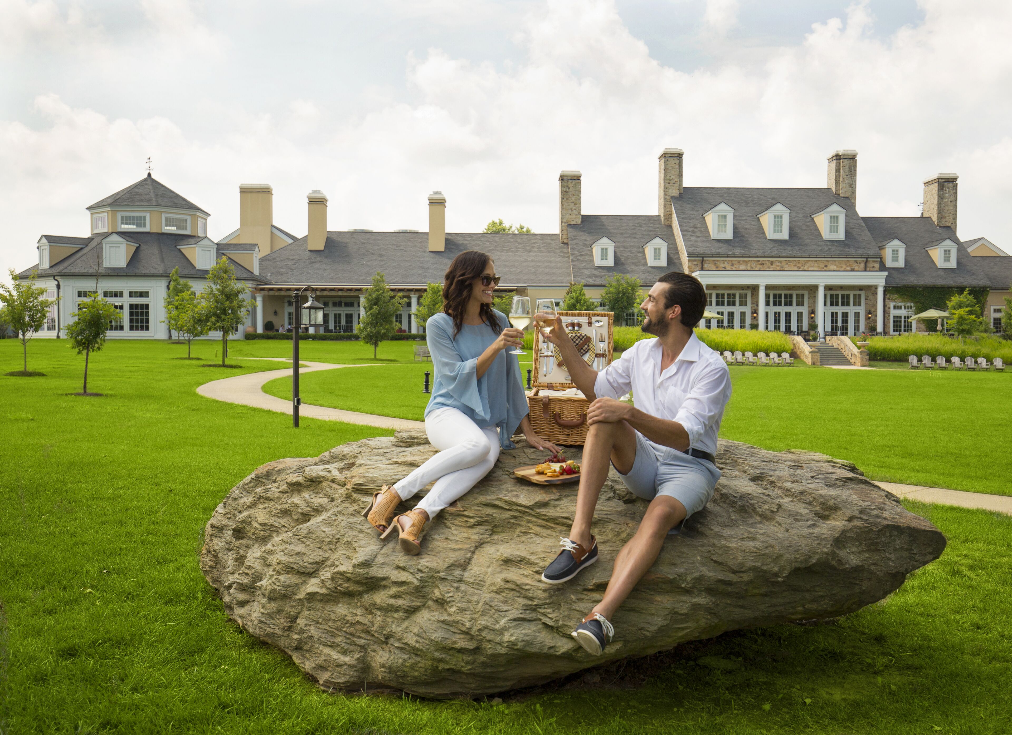 Two people sitting on a rock in a grassy area with a large house in the background.