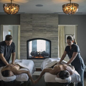 A spa room with two massage tables, a fireplace, and two masseuses attending to clients.