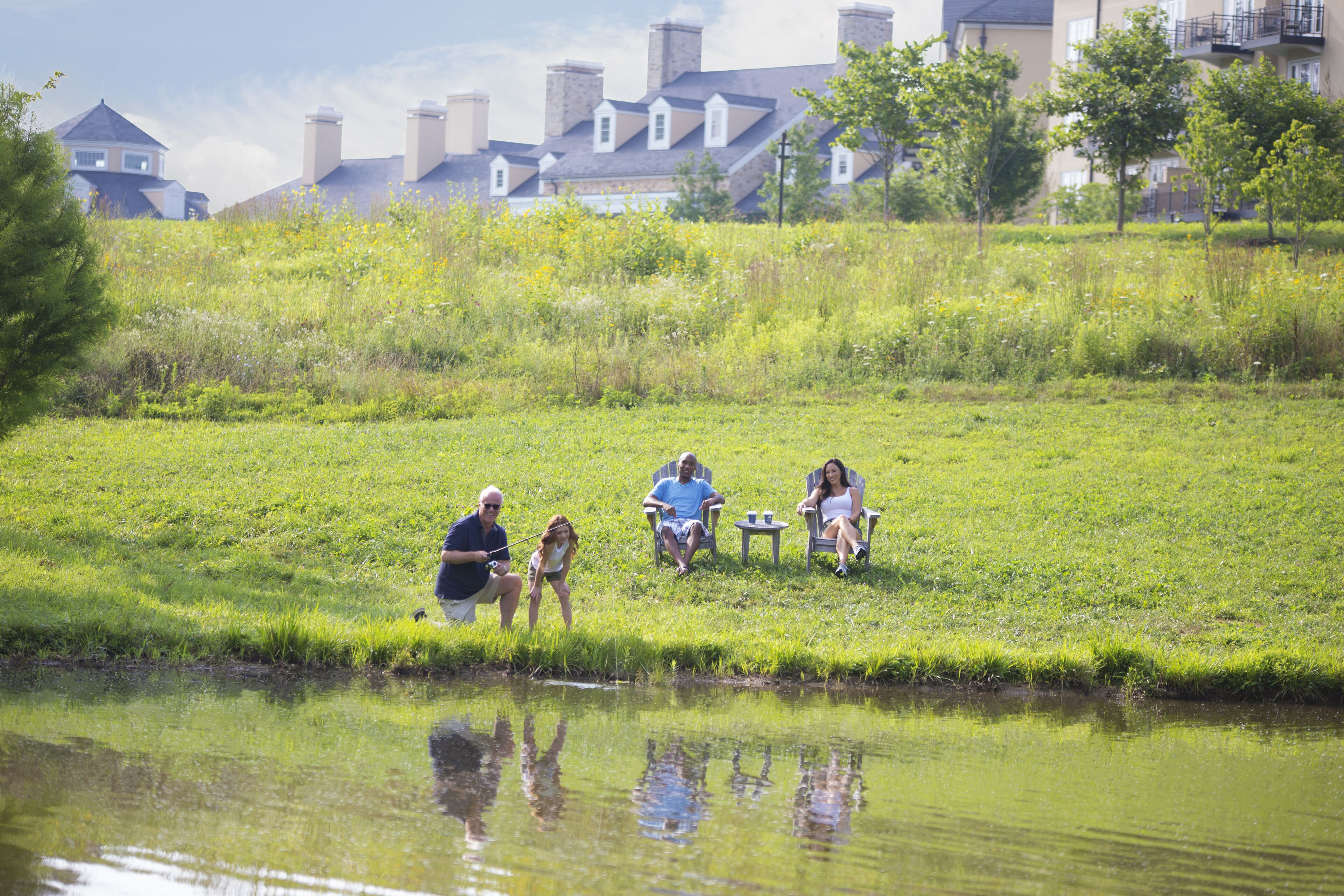 A group of people sitting on chairs by a pond in a grassy area with buildings in the background.