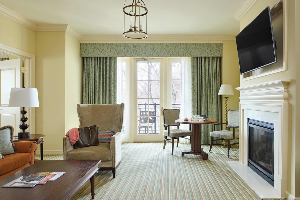 A hotel room with a sofa, armchair, dining table, fireplace, television, and large windows with curtains.