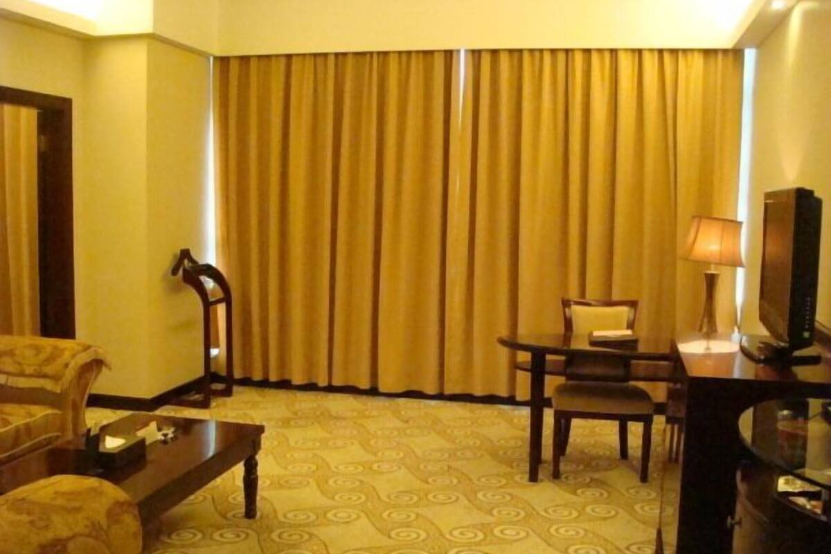 A hotel room with a large window, a desk with a lamp, a chair, a sofa, and a television.
