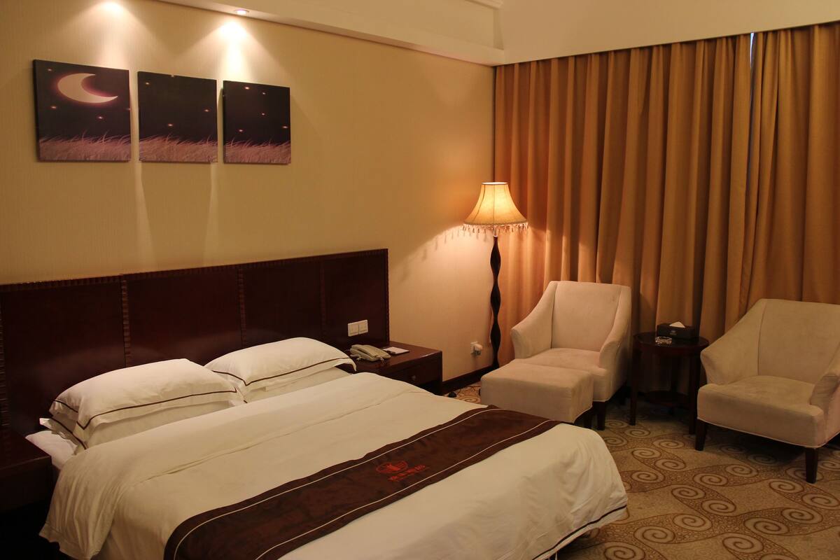 A hotel room with a bed, two chairs, a nightstand, a lamp, and a wall with three paintings.