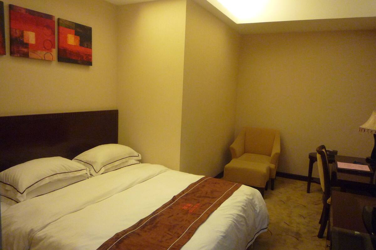 A hotel room with a bed, a chair, a desk, and two paintings on the wall.