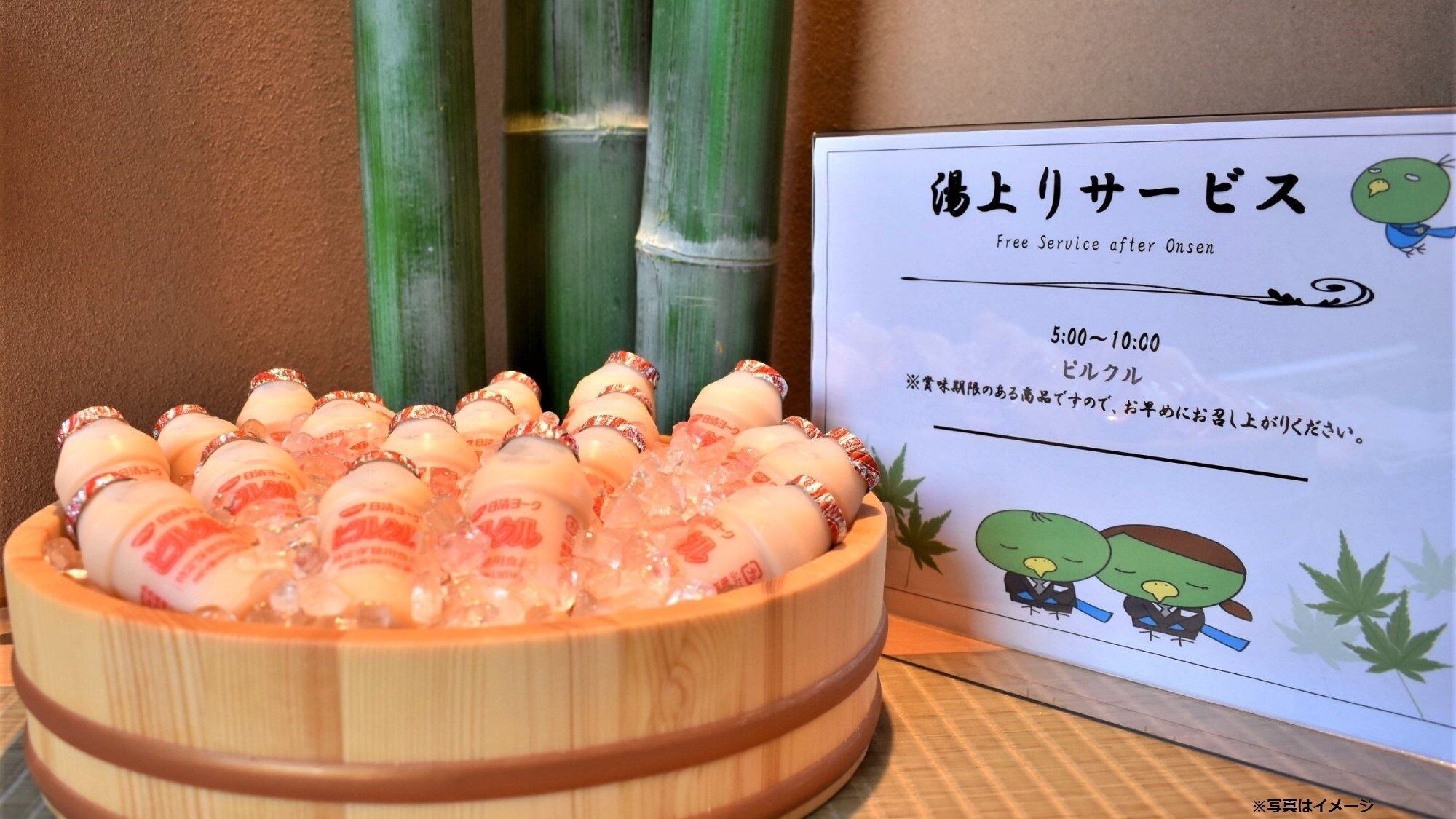 Un bol de fusta ple de botelles de sake, un cartell amb text japonès i plantes de bambú al fons.