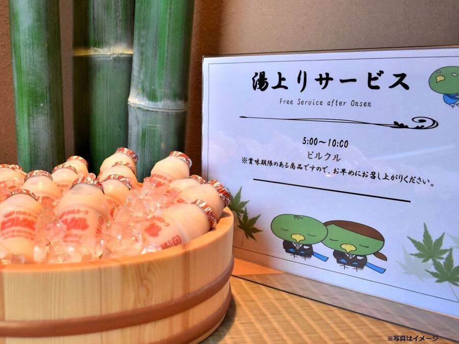 Un bol de fusta ple de botelles de sake, un cartell amb text japonès i plantes de bambú al fons.