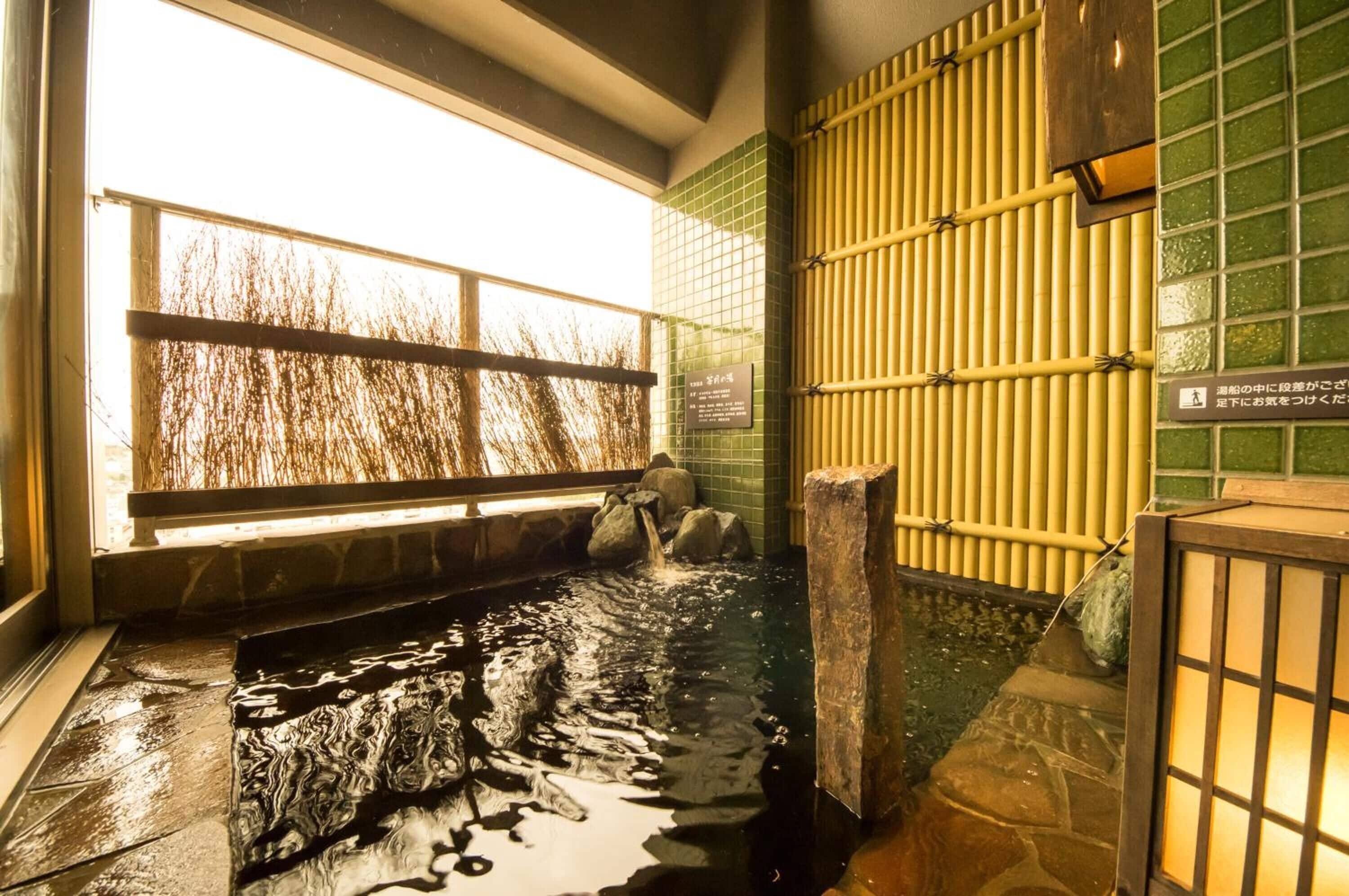 A traditional Japanese onsen with a stone hot spring, bamboo wall, and a view of trees through a window.