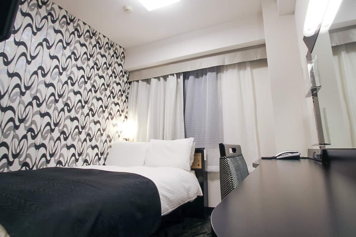 Een hotelkamer met een bed, een bureau, een stoel, een lamp en een raam met gordijnen.