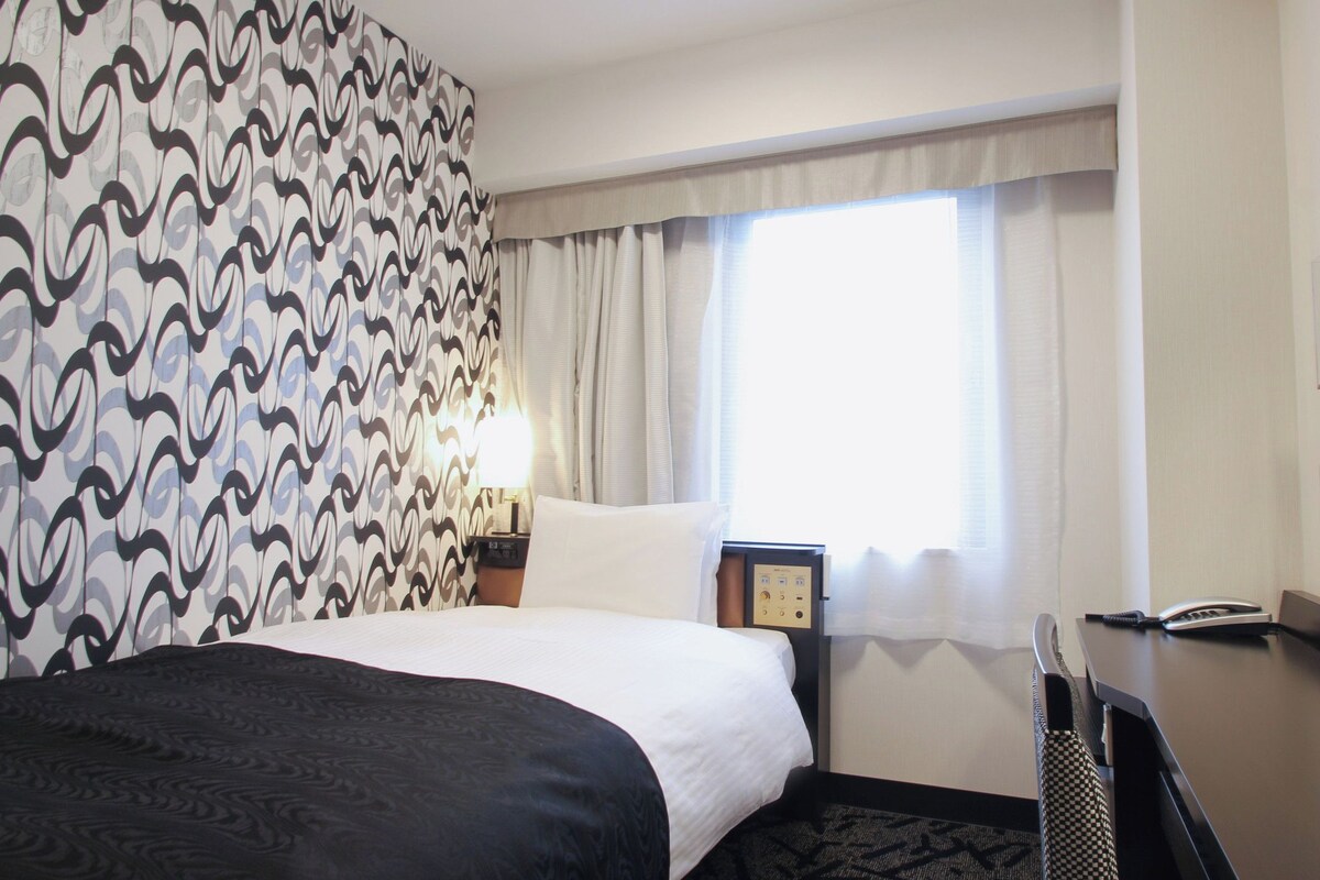 A hotel room with a bed, a desk with a telephone, a mirror, and a window with curtains.