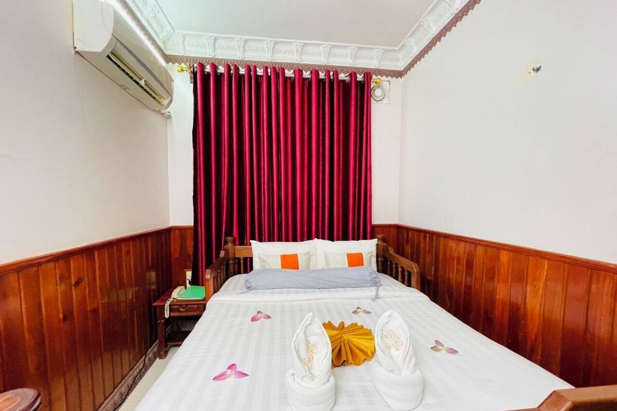 Een hotelkamer met een houten hoofdeinde, een bed met witte lakens, rode gordijnen en een nachtkastje van hout met een groene map.