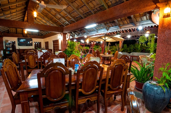A restaurant with wooden tables and chairs, a TV on the wall, and potted plants.