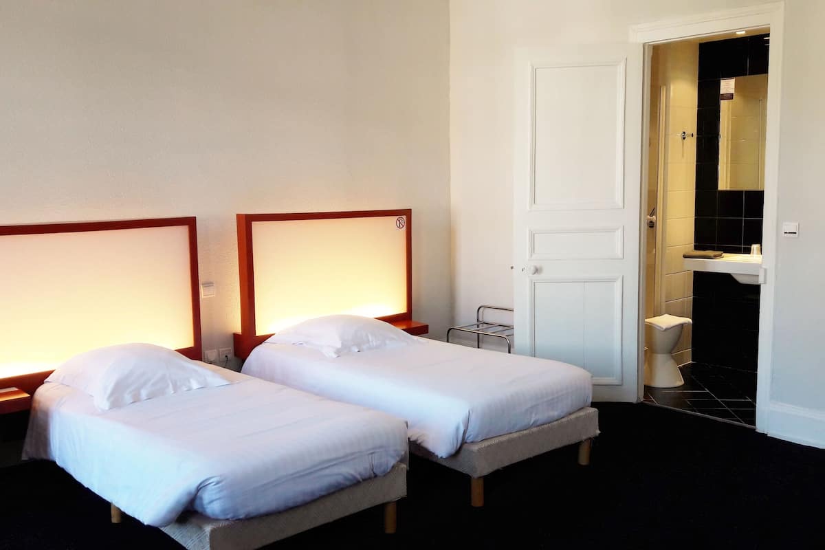 Une chambre d’hôtel avec deux lits simples, une porte blanche et une salle de bain visible, équipée de toilettes et d’un lavabo.