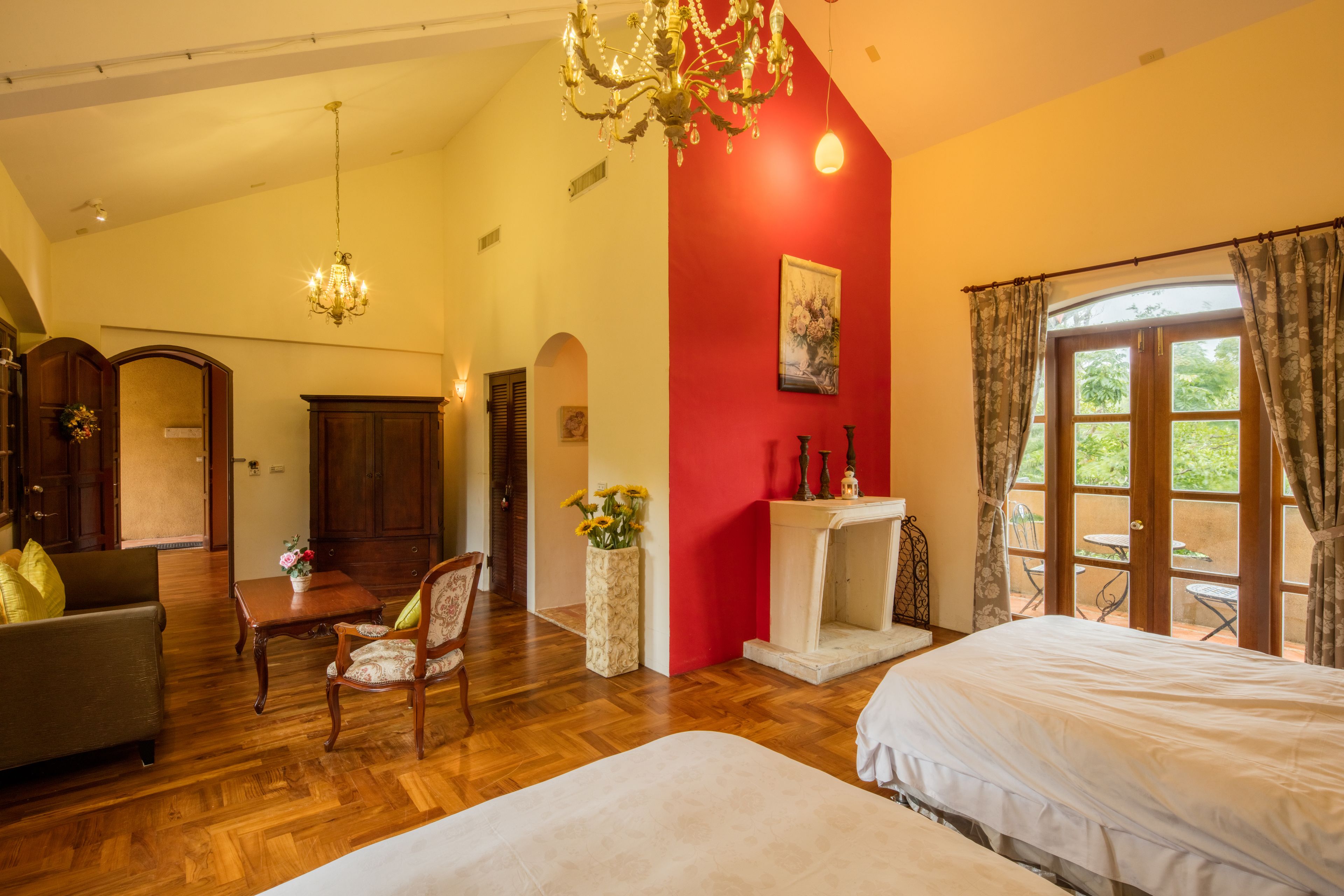 A hotel room with a wooden floor, a red accent wall, a chandelier, a fireplace, and a view through large windows.